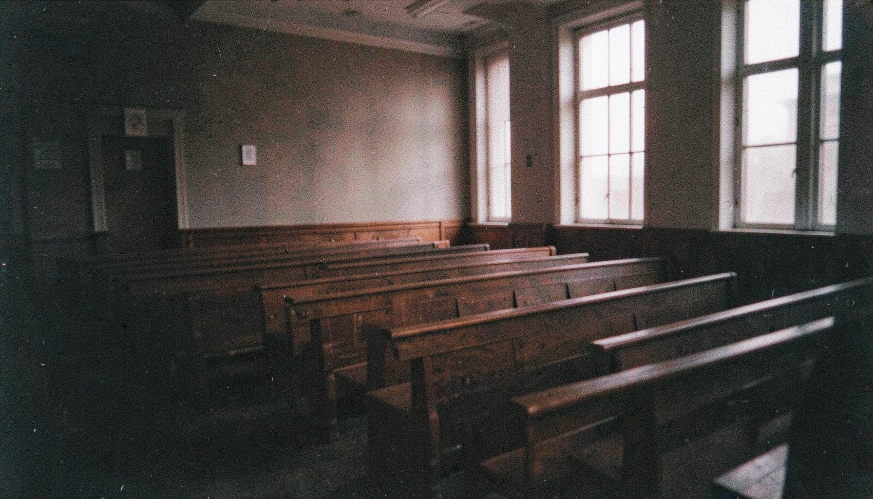 Dark grainy photograph of a quiet courtroom interior with wooden benches and soft light filtering through tall windows, moody and atmospheric
