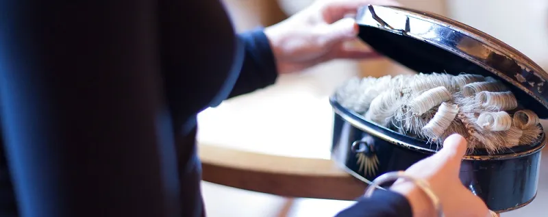 Close-up of hands opening a round tin containing a curled white wig, soft indoor lighting, neutral tones, formal mood.